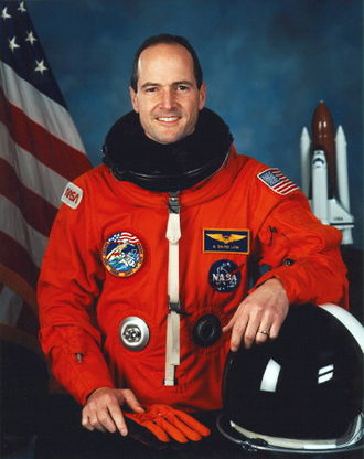 George David Low in astronaut suit stands by U.S. flag